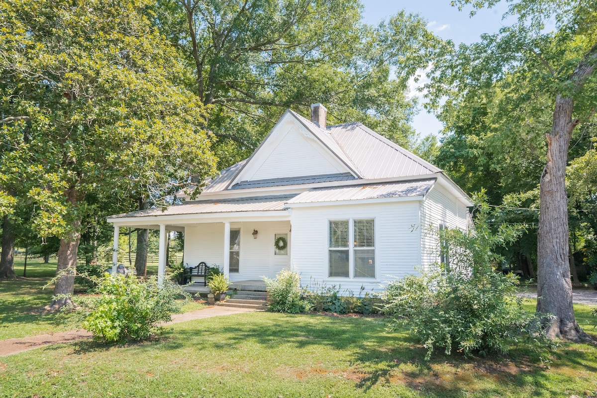 A charming house is framed by lush green trees, highlighting its inviting white exterior. A covered porch with rocking chairs beckons relaxation, while bright windows suggest ample natural light within. The well-maintained lawn and surrounding landscaping enhance the serene country setting.