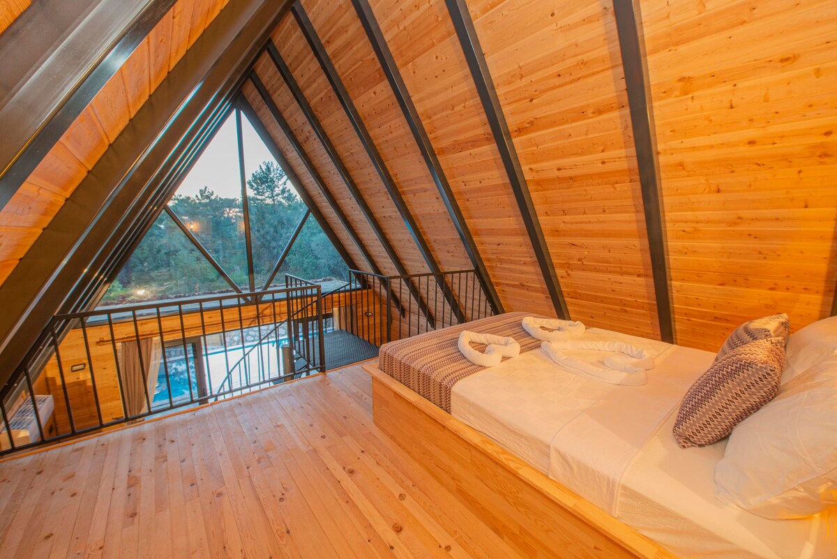 An airy loft space features a spacious bed adorned with neutral-toned textiles. Angled wooden beams rise to meet large triangular windows, offering a view of the surrounding greenery. A wooden railing separates the sleeping area from an open staircase leading to the lower level.