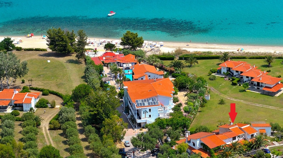 An aerial view showcases a collection of villas with terracotta roofs nestled among lush green gardens. The turquoise sea glistens nearby, with sandy beach visible, and various beachgoers can be seen enjoying the shoreline.