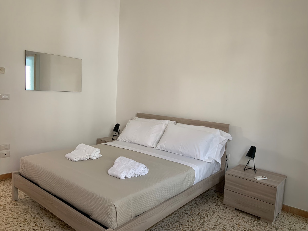 A spacious bedroom features a comfortable bed with neatly arranged white linens and towels. A minimalist wooden frame complements the light-colored walls. A small nightstand with a sleek lamp is positioned on either side of the bed. A large mirror reflects natural light in the room.
