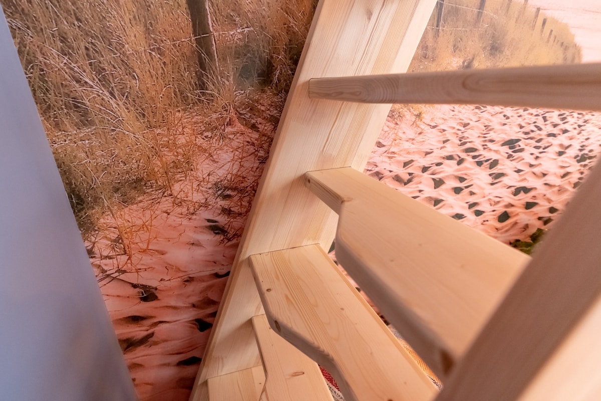 Wooden stairs lead upwards, showcasing smooth natural finishes. The wall behind displays a scenic mural of a sandy beach with tall grasses, enhancing the tranquil atmosphere of the space.