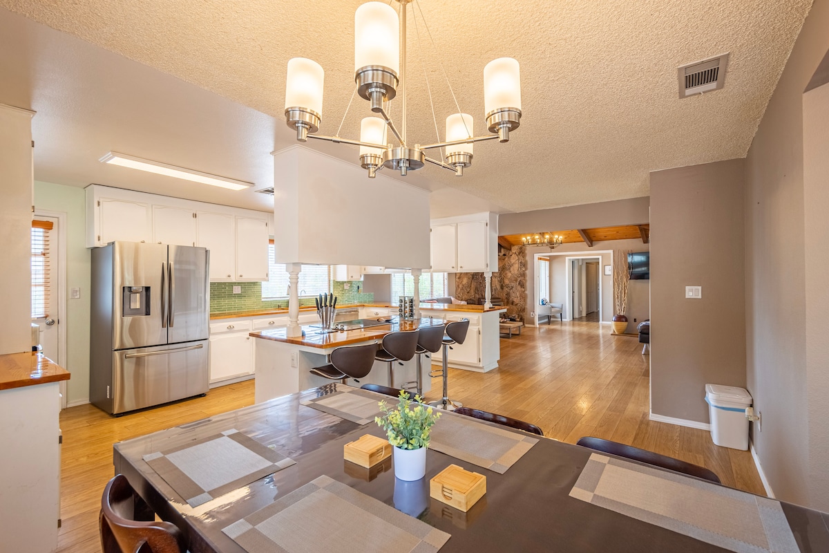 The newly remodeled kitchen features stainless steel appliances and an island bar. A spacious dining area is visible with an 8-seater table, complemented by modern lighting. Natural light enters through adjacent windows, enhancing the inviting ambiance of the open layout.