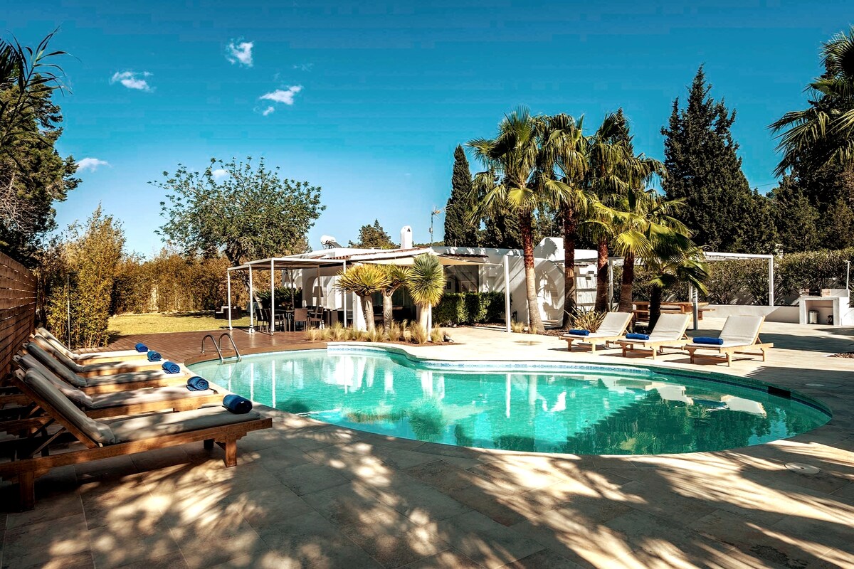 A private pool is surrounded by sunbeds and palm trees, offering a spacious outdoor area with a covered dining space visible in the background. The sky is clear, and the landscape features lush greenery, contributing to a tranquil setting.