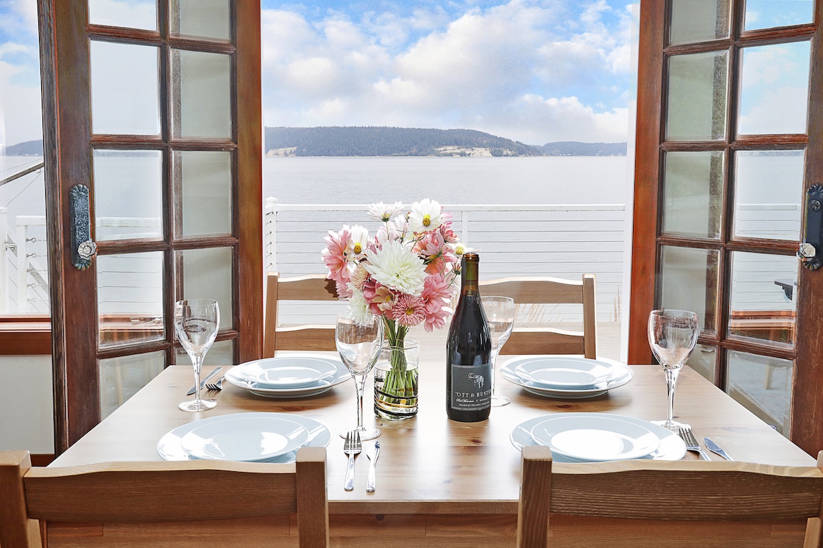 A dining table is set for four, featuring white plates, glassware, and a bottle of wine. A vase of fresh flowers is centered on the table. French doors open to reveal a serene view of the ocean and surrounding landscapes.