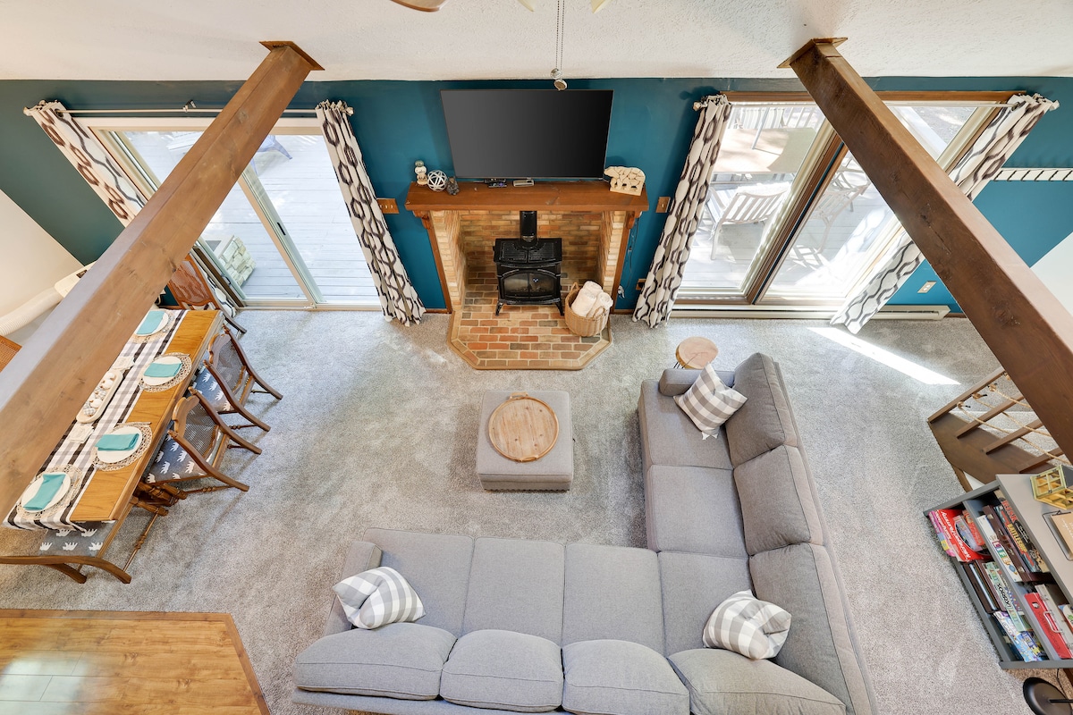 The living area features a large, L-shaped couch positioned around a brick fireplace. Light-colored curtains frame the sliding glass doors, offering views of the outdoor space. A wooden dining table with six chairs is visible, along with various games on a nearby shelf.