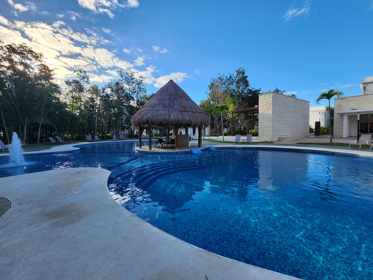 A large pool is surrounded by a spacious deck, featuring a thatched-roof palapa in the center. Lush greenery is visible in the background, contributing to a serene outdoor setting. The water exhibits clear blue tones, reflecting the sky above.