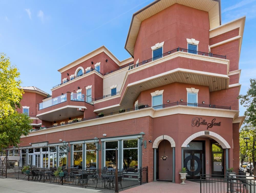 A multi-level building features distinct architectural elements, including a rounded top section, balconies, and large windows. A patio area with black metal furniture is visible, providing outdoor seating. The establishment's name, 'Bella Sant', is prominently displayed near the entrance.