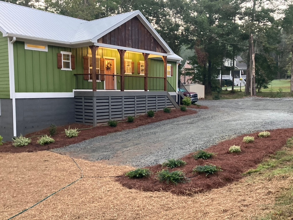 A charming cottage is presented in green and brown hues, showcasing a welcoming front porch with wooden accents. A gravel driveway leads to the property, which features landscaped areas with fresh mulch and shrubs positioned along the front.