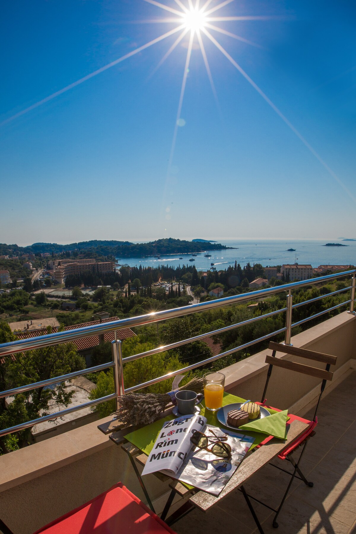 A small outdoor table is positioned on a balcony, offering views of the surrounding landscape and sea. Two red chairs accompany the table, which is set with a decorative green napkin, a piece of cake, sunglasses, and a refreshing drink. Sunlight shines brightly overhead.