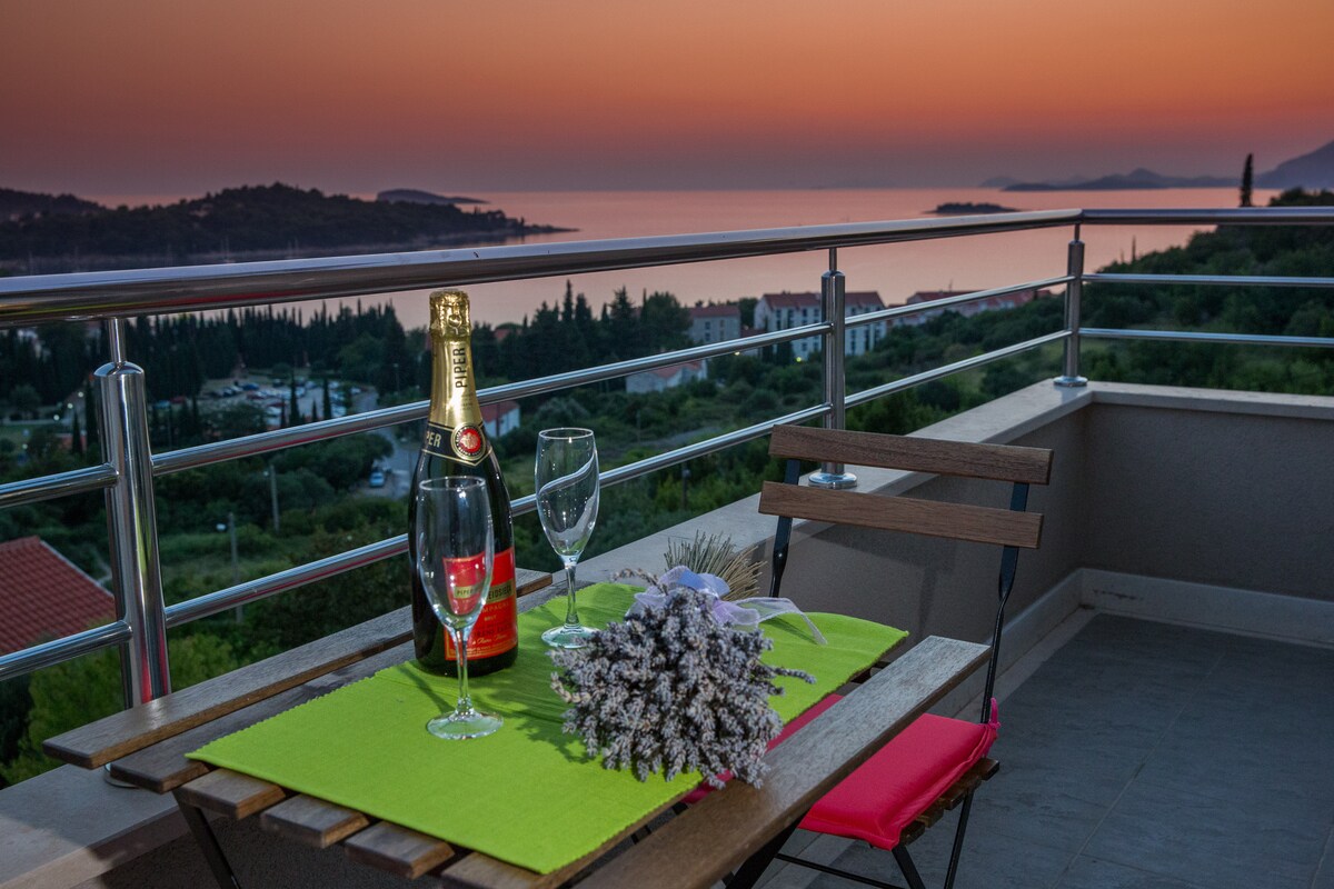 A balcony table is set for two, featuring a bottle of sparkling wine and two glasses. A small bouquet of lavender adds a touch of nature. The background displays a panoramic view of the sunset over the sea, highlighting vibrant oranges and purples in the sky.