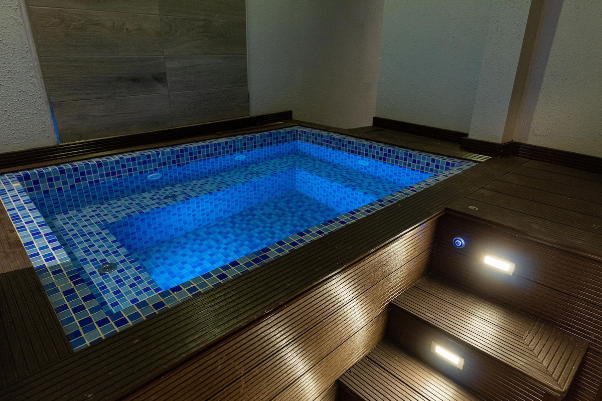 A serene Jacuzzi is surrounded by wooden decking, featuring blue mosaic tiles and soft underwater lighting. Steps lead up from the pool area, providing access to the surrounding space.