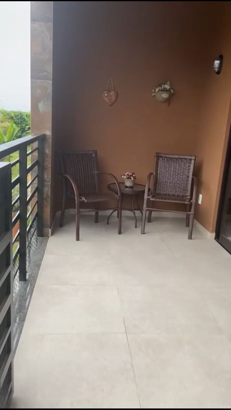 A small outdoor seating area features two woven chairs and a round table set against a warm-colored wall. Decorative wall accents and a potted plant add subtle charm. The space offers a view of the surrounding greenery and is accessible from inside the apartment.