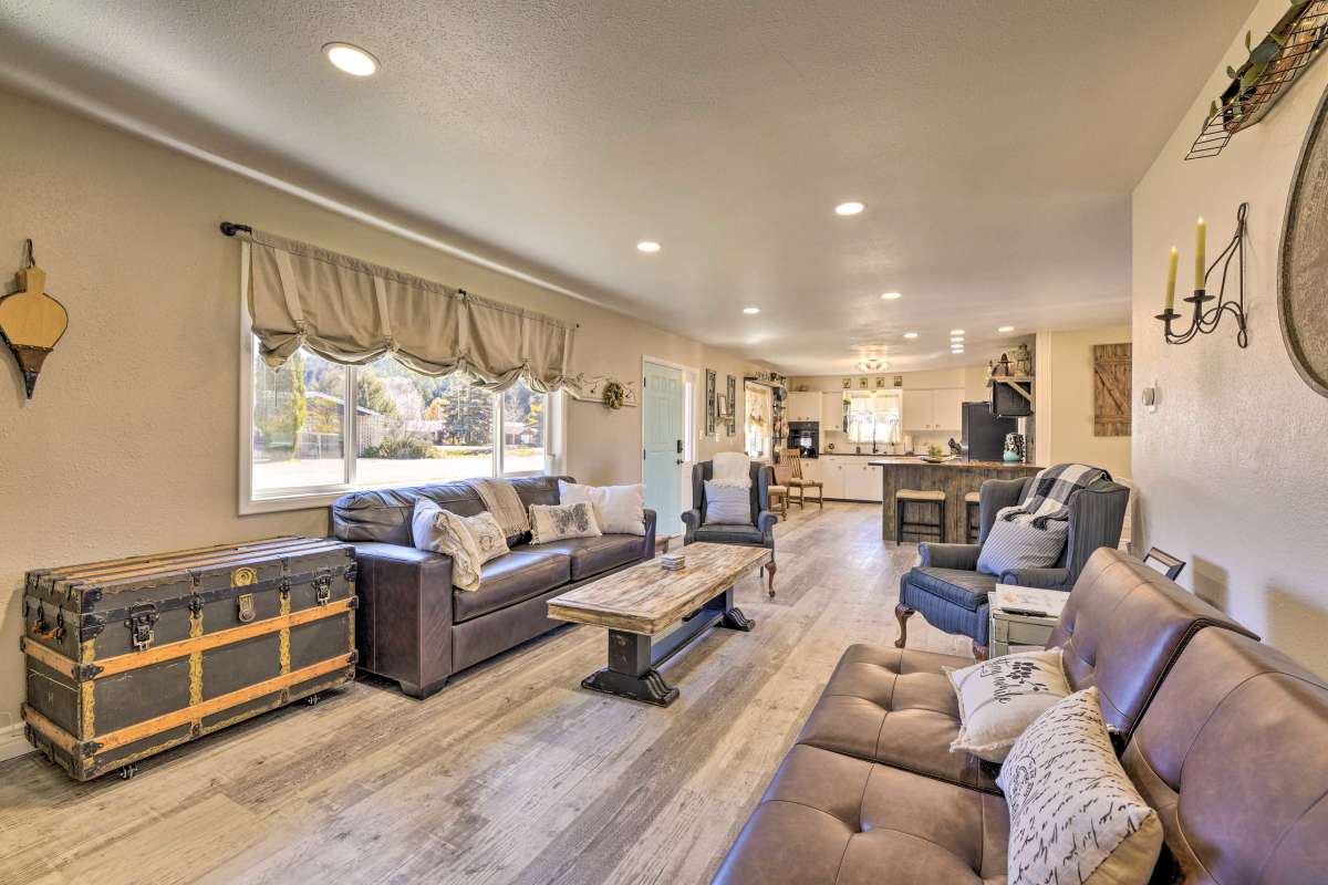 A spacious living area is presented with natural light streaming through large windows. Comfortable seating options include a dark leather sofa and an inviting armchair, complemented by a central wooden coffee table. A vintage trunk adds character to the space, which connects to a modern kitchen area.