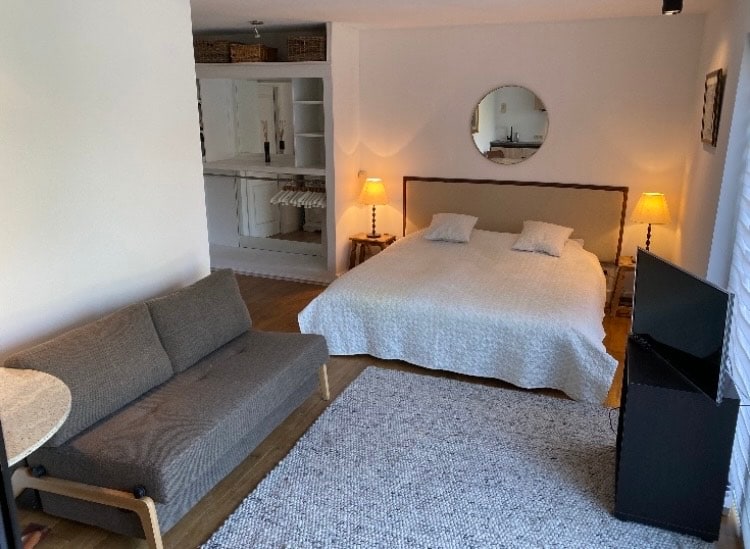 A studio space is presented with a spacious bed dressed in a light coverlet. A comfortable gray sofa and small round table are positioned nearby. Soft lighting is provided by two lamps, and a television stands against the wall. Shelving units are visible in the background.