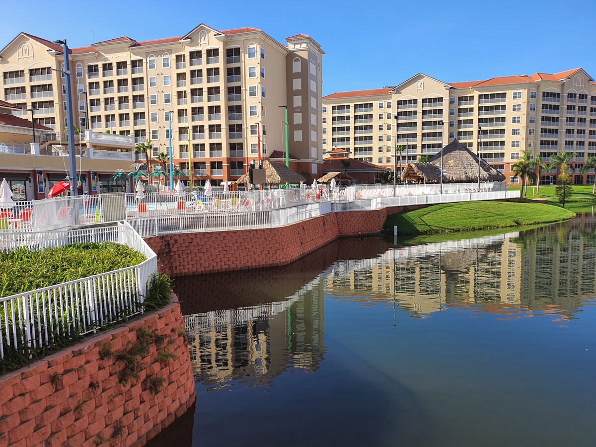 A multi-story resort building stands alongside a landscaped shoreline with a calm body of water reflecting its image. A white fence borders the area, and colorful poolside amenities are visible in the background, suggesting a vibrant atmosphere for relaxation and recreation.
