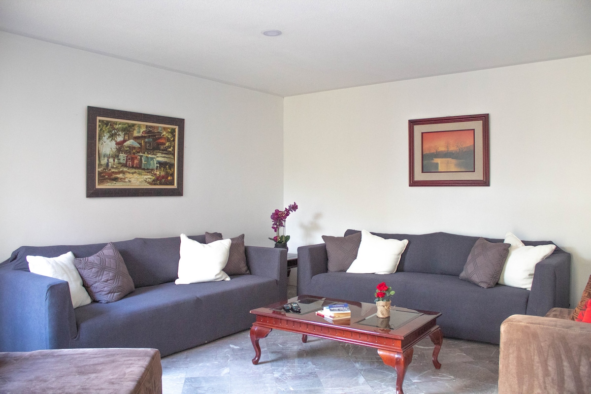 A comfortable living area features two dark gray sofas arranged around a wooden coffee table. Decorative cushions add warmth to the seating. Two framed artworks hang on the wall, and a small flower arrangement sits on the table, enhancing the space's relaxed atmosphere.