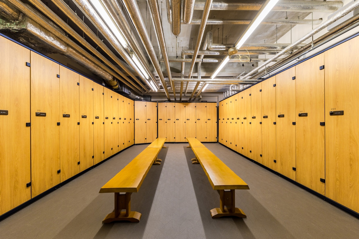 A spacious locker room is depicted, featuring multiple wooden lockers arranged along the walls. Two long wooden benches are positioned centrally, and the ceiling is fitted with exposed piping and bright overhead lighting, creating a functional and well-lit space.