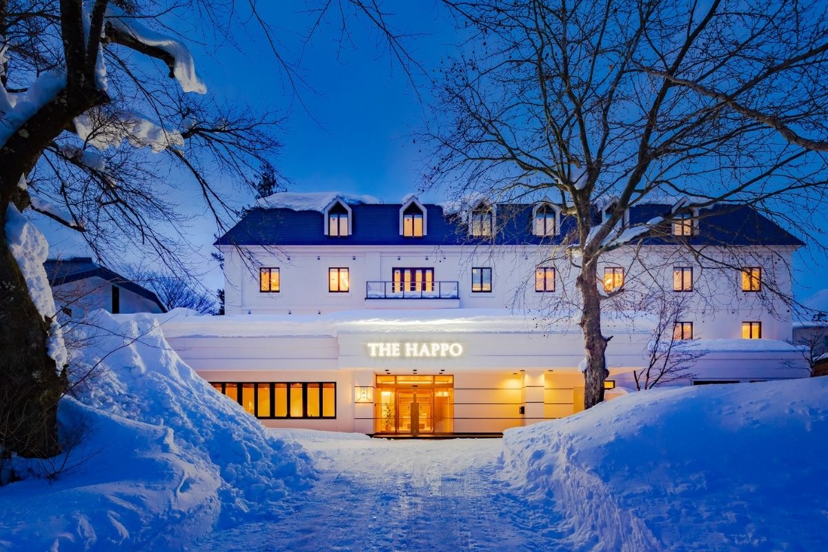 The exterior of The Happo is showcased during twilight, with snow blanketing the ground. Warm lights emanate from the windows, highlighting the sleek design of the building. Trees frame the entrance, creating a serene winter atmosphere.