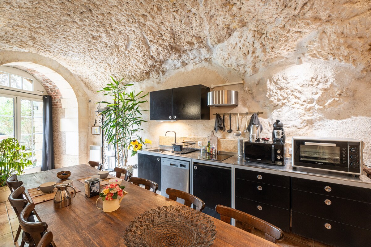 A spacious kitchen features a long wooden dining table set with a decorative centerpiece. Black cabinetry contrasts against the natural stone walls, highlighting modern appliances such as a microwave, coffee maker, and sink. Large windows provide natural light and views of the surrounding greenery.