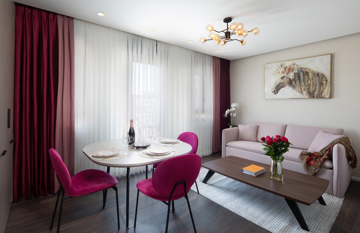 The living area features a round dining table set for four, surrounded by pink chairs. A modern couch sits adjacent, complemented by a table with decorative items. Bright curtains frame the windows, allowing natural light to fill the space, while wall art adds a subtle touch.