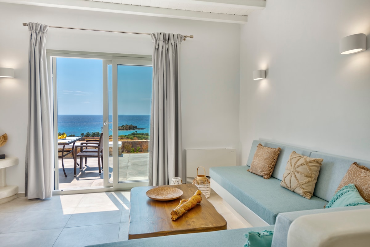 A bright living area features light blue sofas adorned with decorative pillows. Large glass doors provide access to an outdoor space with a view of the Aegean Sea. Natural light fills the room, and a wooden coffee table is positioned centrally.