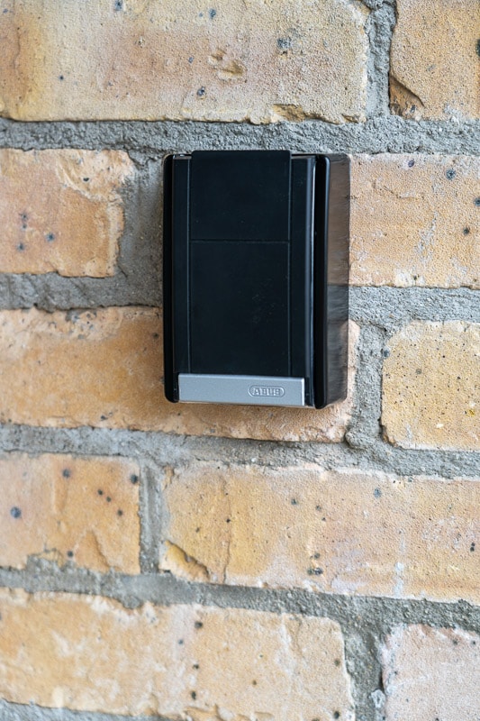 A black keypad is mounted on a brick wall, featuring a smooth surface and a small silver area at the bottom. The surrounding bricks are a warm, light brown, showcasing a subtle texture and color variation.