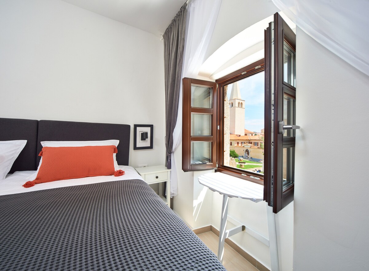 A cozy sleeping area features a spacious bed adorned with neutral bedding and a vibrant orange accent pillow. An arched window opens to reveal scenic views, allowing natural light to brighten the room. A small table rests beside the bed, enhancing the functional layout.