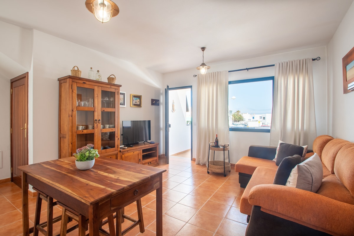 A bright living area features a wooden table surrounded by chairs. A comfortable orange sofa is positioned near large windows, allowing natural light to fill the space. A wooden cabinet holds decorative items, while a television stands nearby, enhancing the cozy atmosphere.