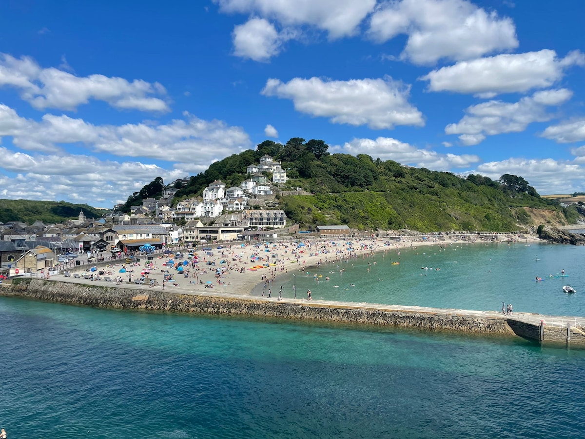 A wide view of a beach reveals golden sand filled with sunbathers and families enjoying their day. The coastline features lush green hills leading up to a residential area, with scattered white buildings visible in the background under a blue sky dotted with fluffy clouds.