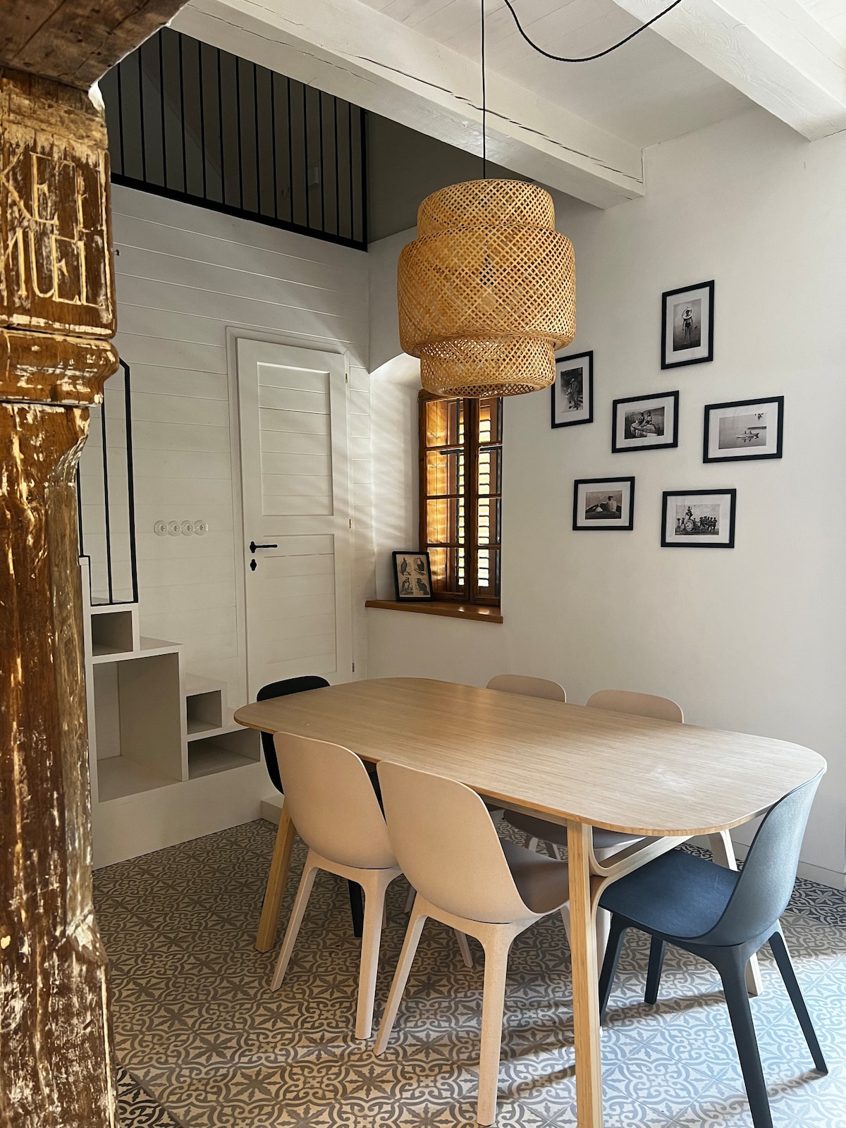 A modern dining area features a wooden table surrounded by six chairs in neutral tones. A woven pendant light hangs above, complemented by wall art displayed in black frames. Natural light fills the space through a nearby window with wooden shutters.