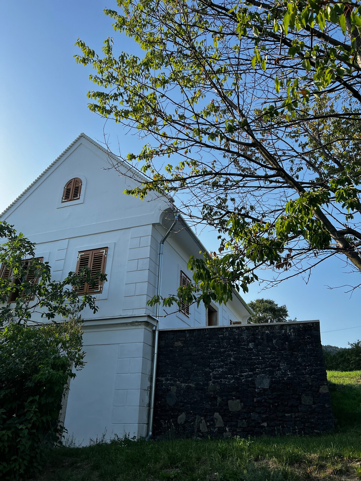 The exterior of the house is shown, highlighting its white walls and unique architectural features, including shuttered windows. A tree is visible nearby, complementing the natural surroundings of the property, while a stone wall provides a structured boundary.