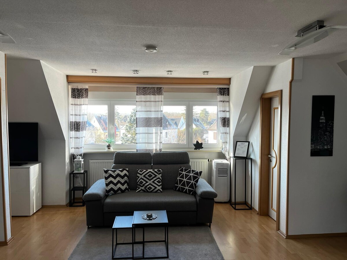 A modern living area features a gray sofa adorned with black and white cushions. A small coffee table is centered on a soft rug, while large windows let in natural light, framed by sheer and patterned curtains. A flat-screen TV is positioned against one wall.