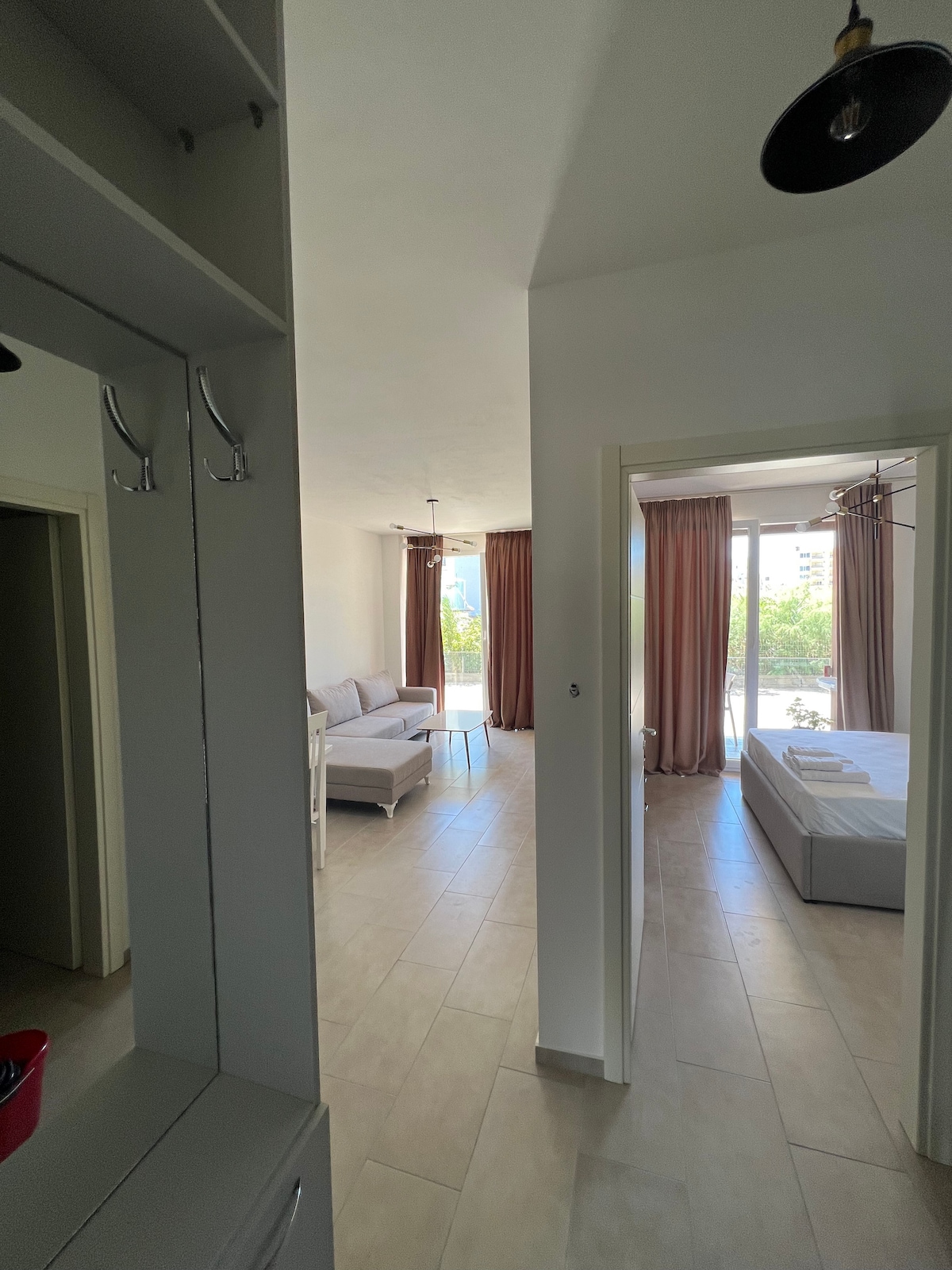 A well-lit interior space is captured, showcasing a living area with a light-colored sofa and large windows framed by soft curtains. A spacious bed is visible in the background, accompanied by a neatly arranged set of linens. Smooth, tiled flooring enhances the open feel of the area.