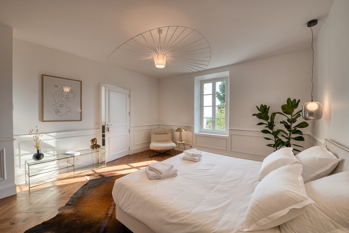 The spacious bedroom features a large bed with white linens, complemented by soft natural light filtered through a nearby window. A minimalist artwork adorns the wall, while a green plant and a modern lamp enhance the room's serene atmosphere.