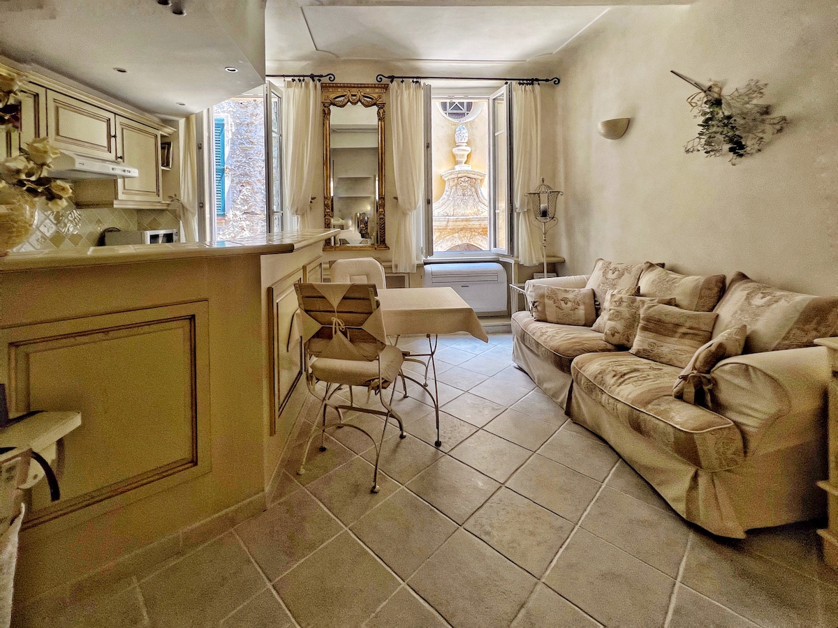 A cozy interior space features a light-colored sofa and a small dining table. A well-equipped kitchen area is visible, with cabinetry and appliances. Natural light enters through a large window, framed by soft curtains, illuminating the tiled floor.