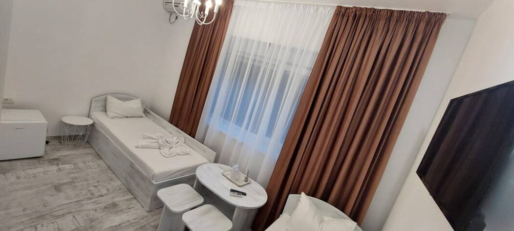 A bright room features a spacious bed with fresh linens and a cozy seating area. Light curtains frame the window, allowing natural light to fill the space. A round table is surrounded by three chairs, and a wall-mounted television adds convenience.