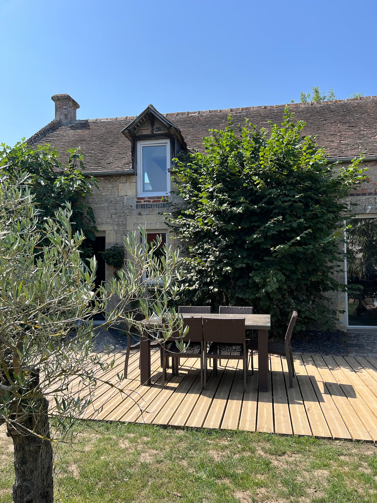 An outdoor wooden terrace features a dining table and chairs, surrounded by lush greenery. The stone house, with a sloped roof and large windows, is partially obscured by a leafy tree, providing a sense of privacy and nature.