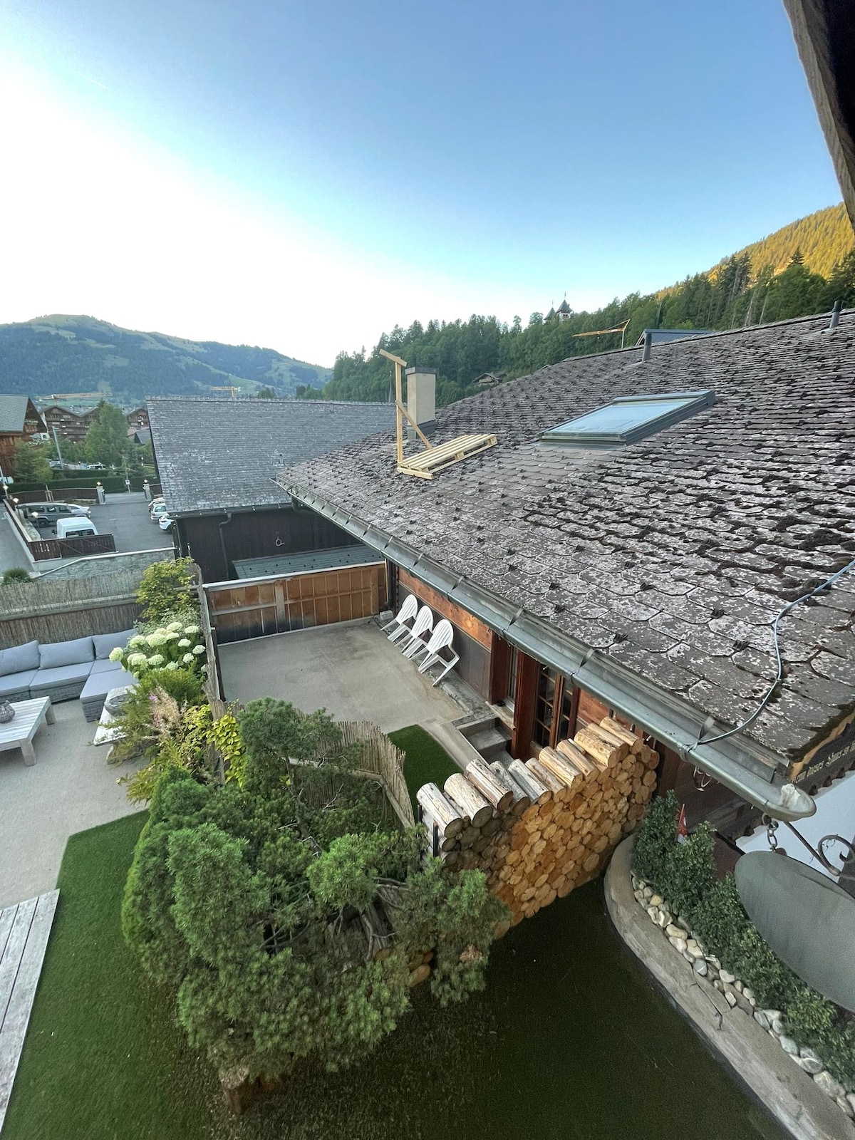 An aerial view shows a rustic chalet featuring a sloped roof with weathered shingles. A neat landscape includes a small garden with greenery and stacked firewood. An outdoor seating area is visible with white chairs positioned on a stone patio.