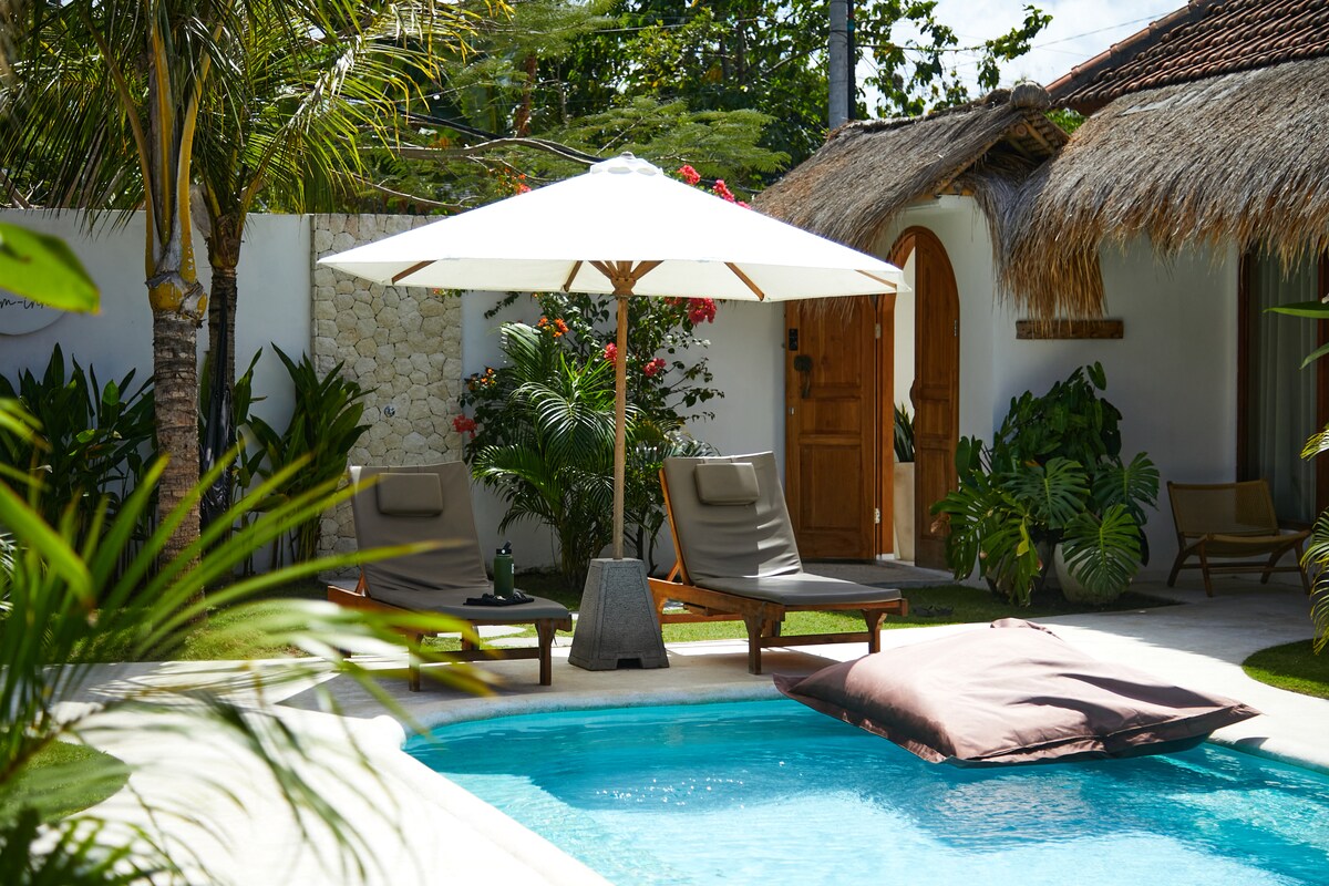 A serene pool area is highlighted by a sparkling blue pool surrounded by lush greenery. Two comfortable lounge chairs are placed beneath a large white umbrella. A cozy bean bag rests against the pool's edge, inviting relaxation.