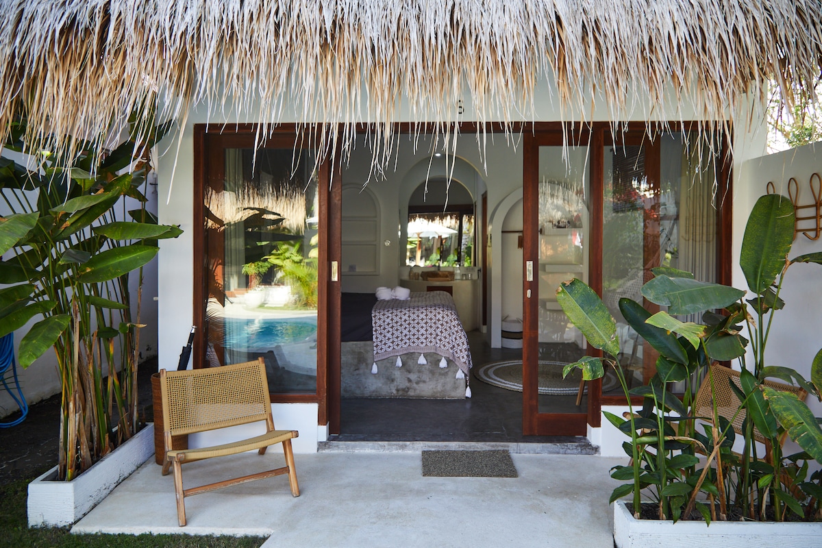 A well-appointed room features large glass doors that open to a private outdoor area. Lush greenery surrounds the entrance, including tropical plants. Inside, a spacious bed is visible, paired with neutral decor, creating a serene environment. A cozy seating option is placed on the patio.