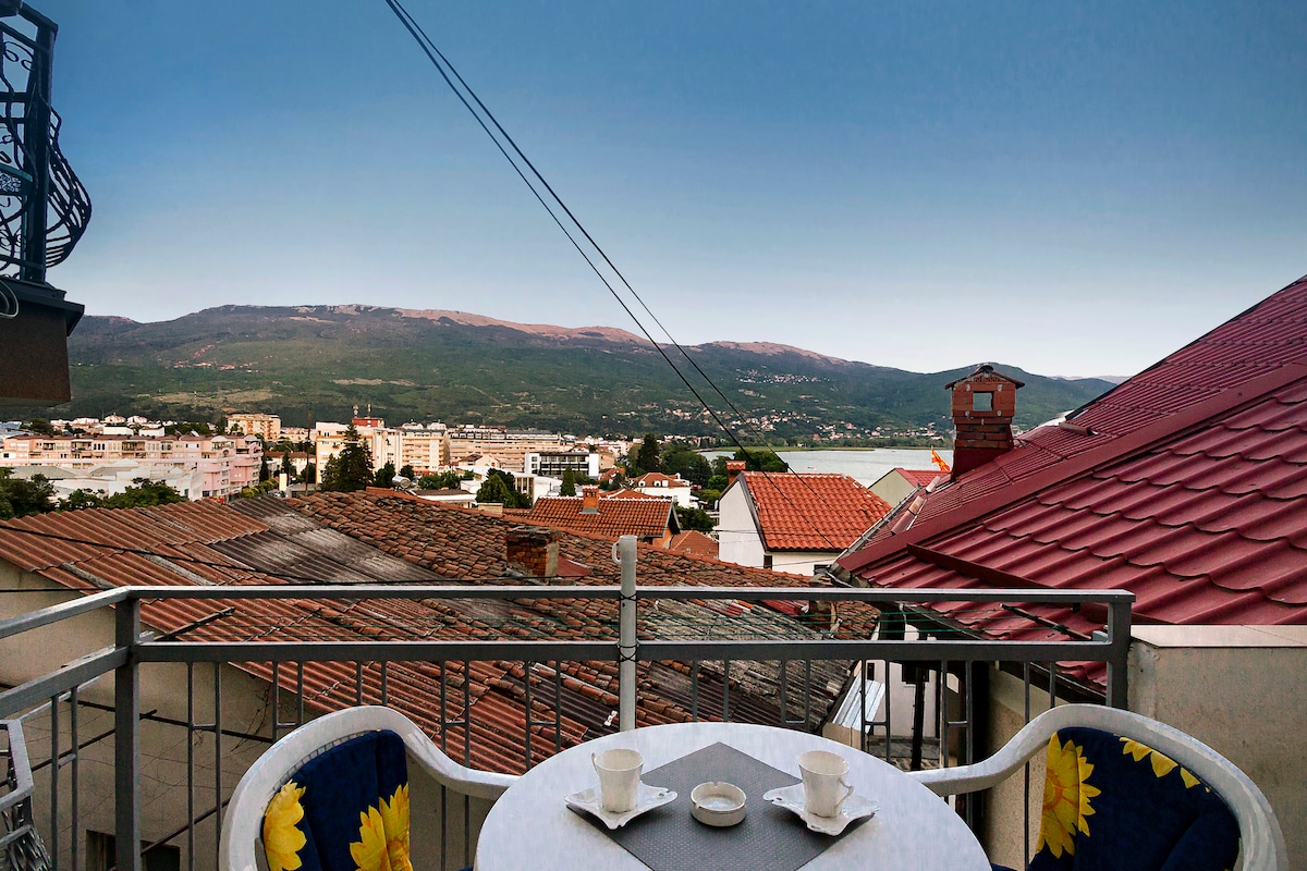 A balcony view reveals a scenic landscape of mountains and town rooftops. Two small cups are placed on a round table adorned with a grey cloth. The surrounding area features a mixture of tiled and flat rooftops under a clear sky.