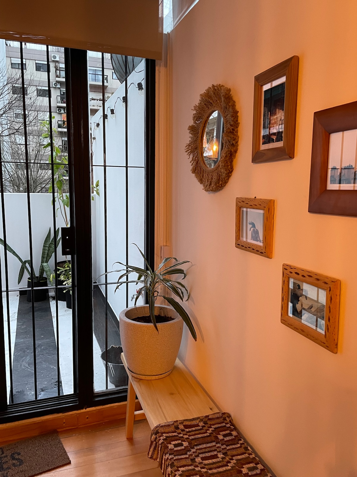 A welcoming entry area features a wooden bench beside potted greenery. Framed photographs are displayed on the wall, alongside a round mirror. Light influxes through large glass doors, providing a view of the terrace, while a textured rug adds warmth to the wooden flooring.