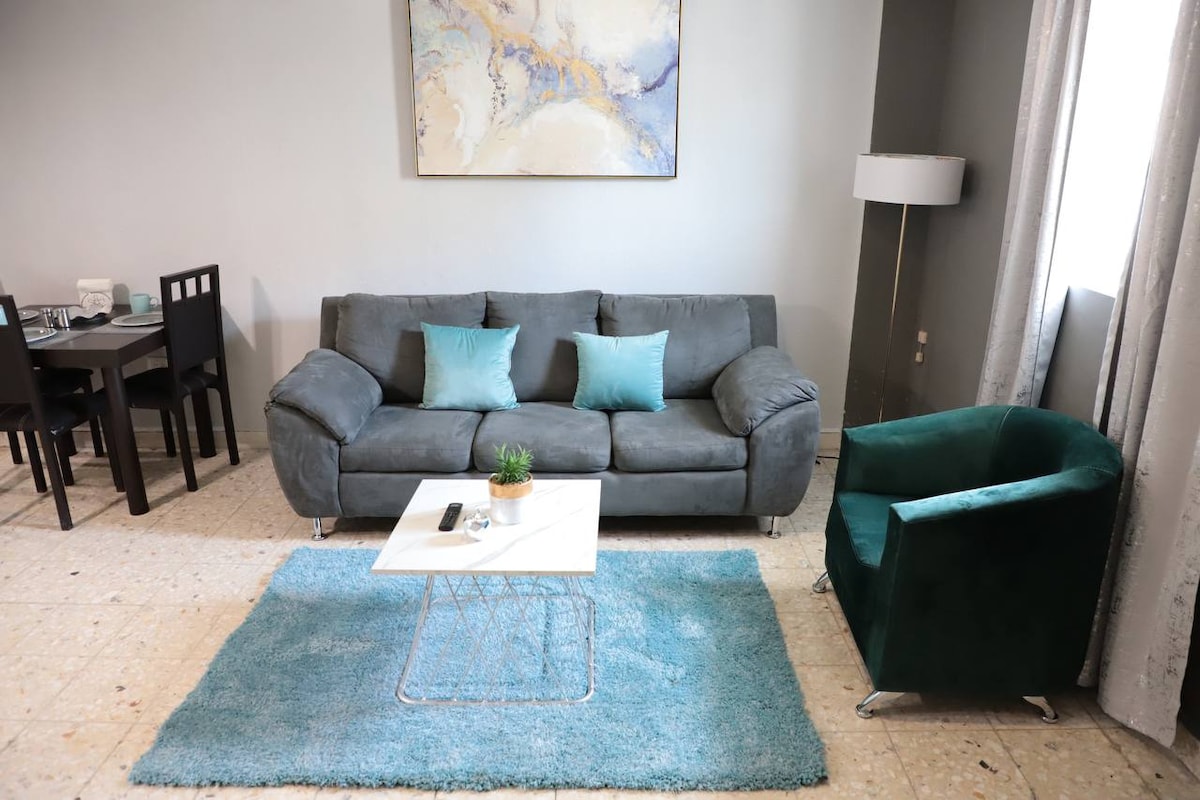 A comfortable living area is displayed, featuring a gray sofa adorned with teal cushions, accompanied by a green armchair. A glass coffee table sits on a light blue rug, and a piece of abstract wall art adds color above. Natural light filters through sheer curtains.