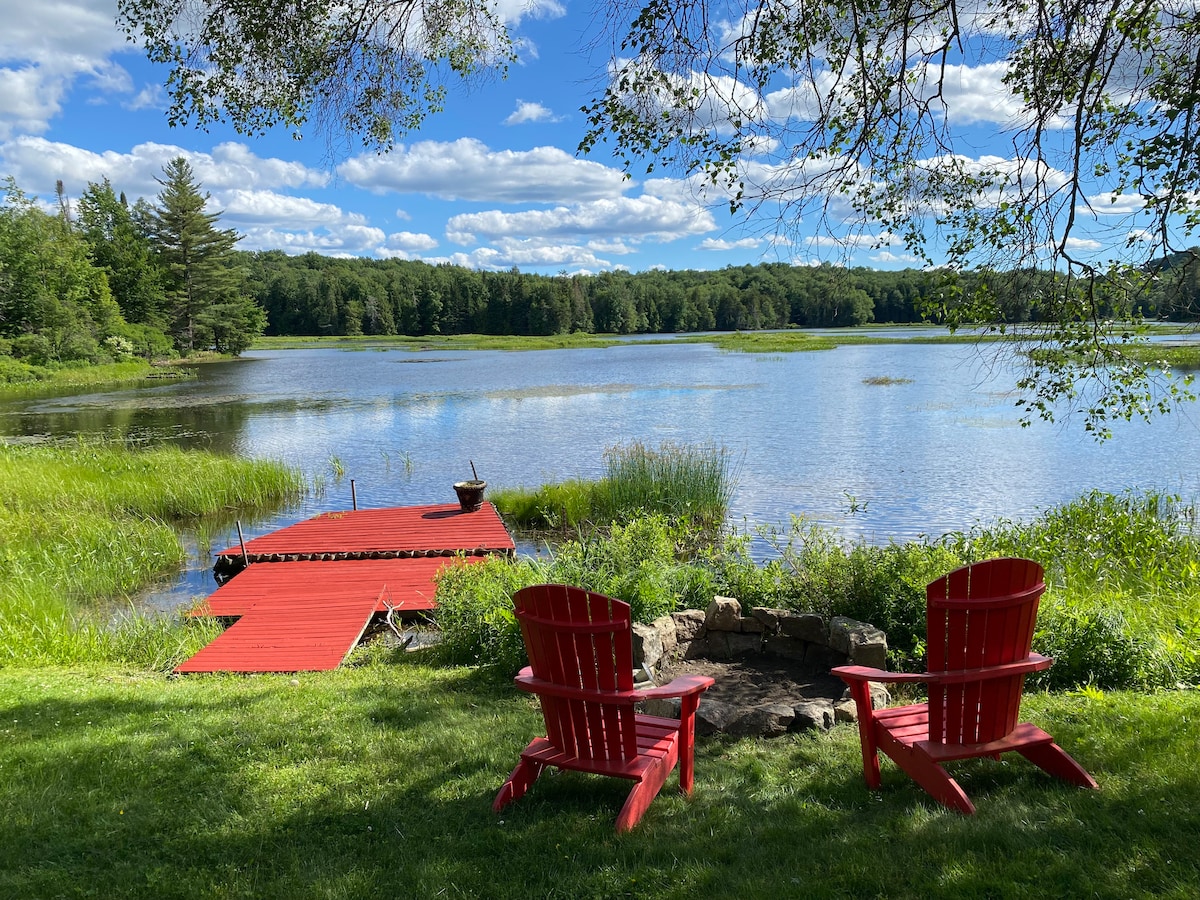 Two red Adirondack chairs are positioned near a fire pit, facing a serene waterfront view. A wooden dock extends into the water, surrounded by lush green grass and natural scenery, creating a peaceful outdoor space.