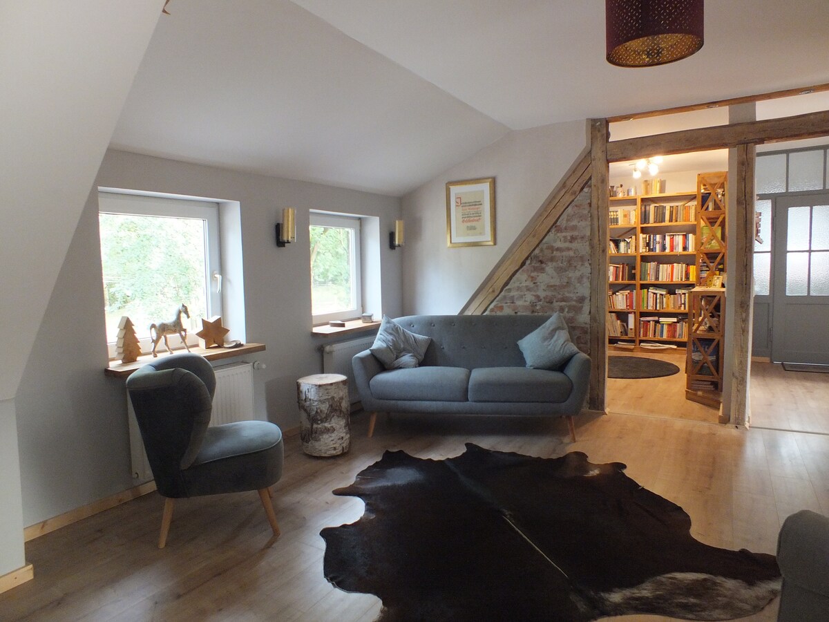A cozy living area features a gray sofa and a rounded chair, arranged atop a natural-toned cowhide rug. Warm light is provided by lamps mounted on the walls, while windows allow for views of the surrounding greenery. Rustic wooden accents and a bookshelf are visible in the background.