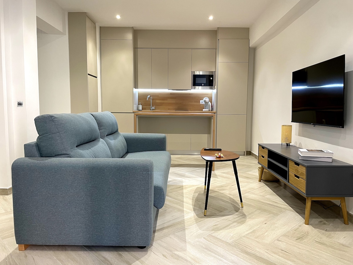 A contemporary living area features a comfortable light blue sofa in front of a medium-sized wooden coffee table. A wall-mounted TV is positioned above a sleek media unit. The kitchen area, with modern cabinetry and appliances, is visible in the background.
