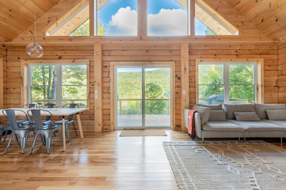 Bright and spacious interior features wooden walls and large windows that offer scenic mountain views. A dining area with a table is visible alongside a cozy living space with a couch. Natural light fills the room, enhancing the inviting atmosphere.