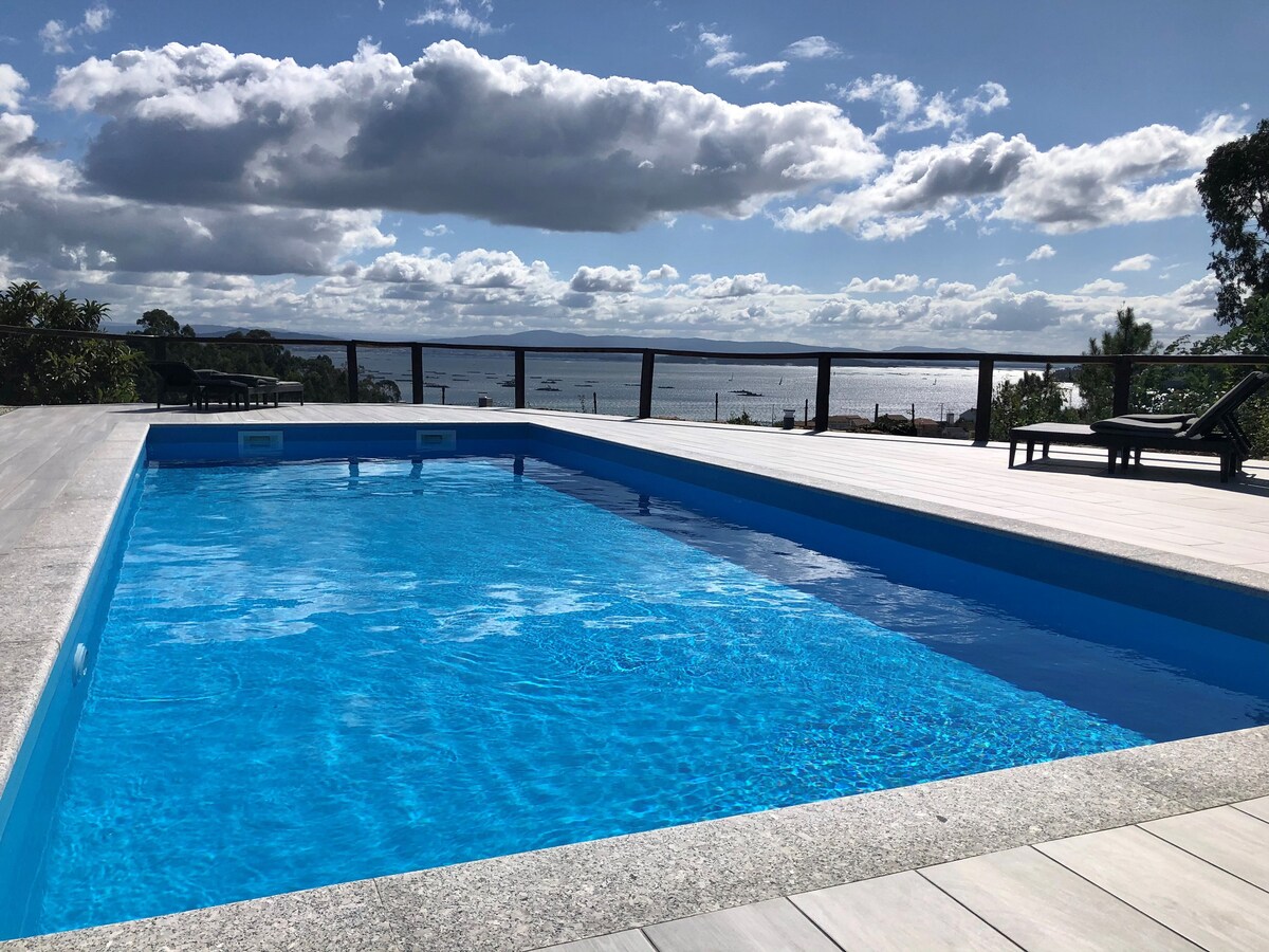 An infinity pool glistens under a clear blue sky, with gentle clouds reflecting above. The pool's edge seamlessly merges with views of the ría de Arousa in the distance. Surrounding lounge chairs provide a relaxing spot to enjoy the serene outdoor setting.