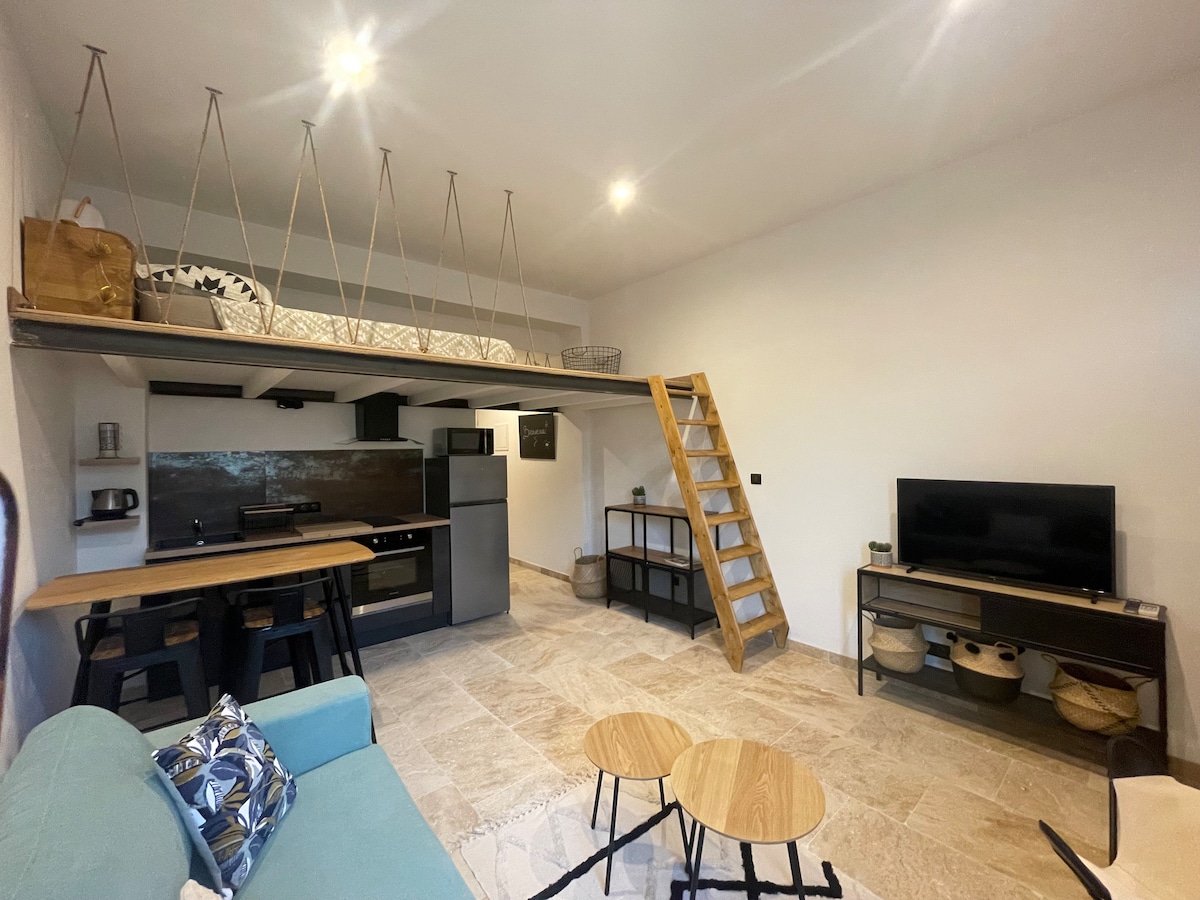 An open living area is featured with a kitchenette, dining table, and cozy seating. A small staircase leads to a lofted sleeping area. Two wooden side tables are positioned nearby, and a television is mounted on a shelf against the wall.