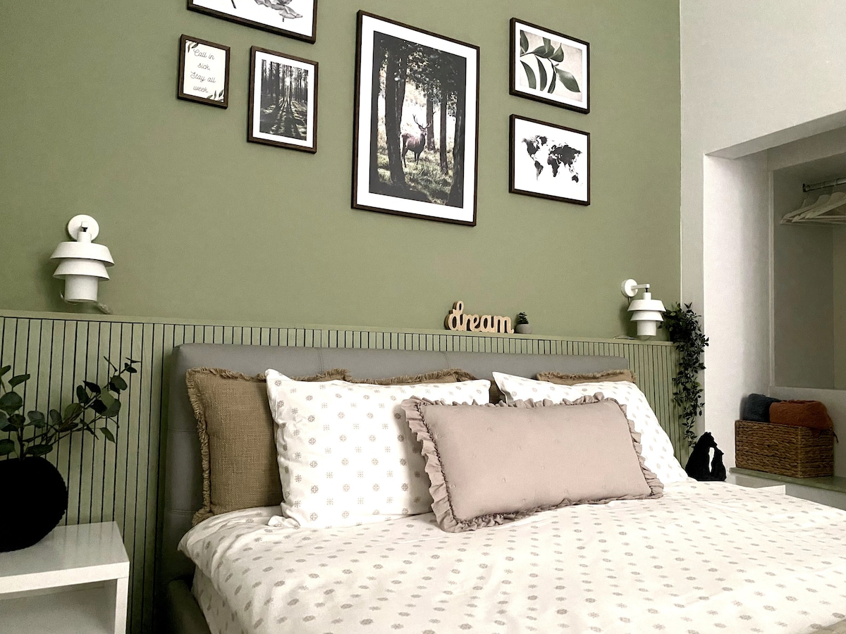 The bedroom features a spacious bed adorned with patterned bedding and decorative pillows. Green accent walls are complemented by framed prints and wall decor. Soft lighting is provided by bedside lamps, with plants adding a touch of nature to the inviting space.